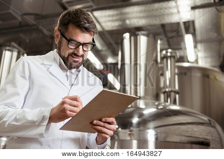Joyful and satisficed. Smiling glad young man making notes at beer factory while working and spending time in the machinery.