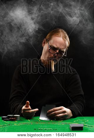 Young red-haired bearded man in glasses and black pullover with a cigar and cards sitting at poker table covered with green cloth in a puff of smoke.