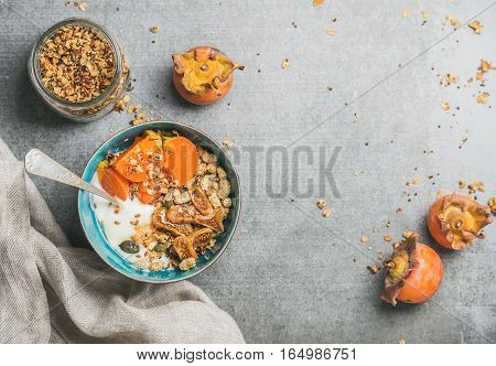 Healthy vegetarian breakfast. Oatmeal, quinoa granola with yogurt, dried fruit, seeds, honey, fresh persimmon in blue bowl over grey background, top view, copy space. Dieting, allergy-friendly concept