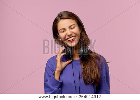 Beautiful Brunette Girl Laughs And Smiles Happily Closing Her Eyes. She Holds In Her Hand The Headph