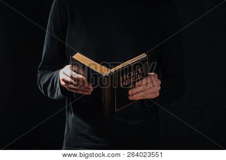 Cropped Shot Of Priest In Black Cassock Holding Holy Bible Isolated On Black
