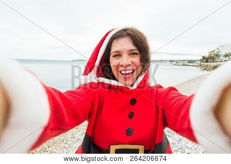 Christmas, Humor And People Concept - Young Woman In Santa Suit Making Selfie Near The Sea At Summer
