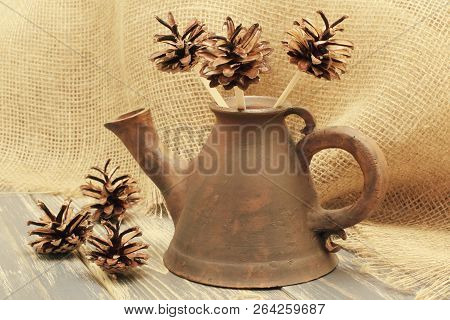 Exclusive Clay Teapot Of Unusual Shape With Decorative Flowers From Open Pine Cones On The Backgroun