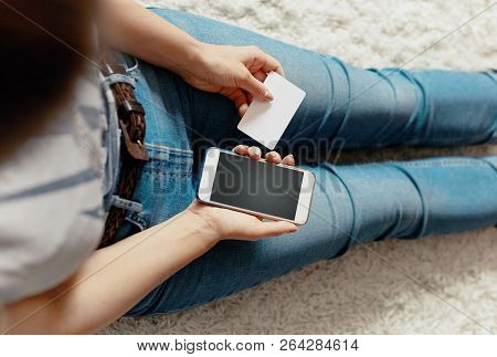 Young Woman Holding Credit Card And Using Phone Sitting On Floor. Payment.  Online Shopping Concept.