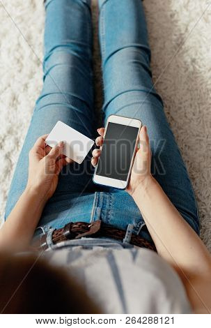 Young Woman Holding Credit Card And Using Phone Sitting On Floor. Payment.  Online Shopping Concept.