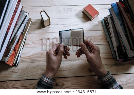 A Man Reading A Miniature Book Among The Big Books In The Library At The Wooden Table.