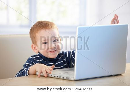 ¿Niño feliz jugando con el ordenador portátil, sentado en la mesa, sonriendo.?
