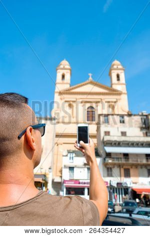 closeup of a young caucasian man, wearing sunglasses, taking a picture of the Saint-Jean-Baptiste Church in the old town of Bastia, in Corsica, France, with his smartphone
