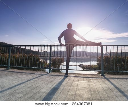 Runner Doing Stretching Exercise On Bay Bridge. An Active Wiry Man Standing On One Legs And Stretchi