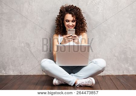 Happy Young Woman Sitting On The Floor With Crossed Legs And Using Laptop On Gray Background.