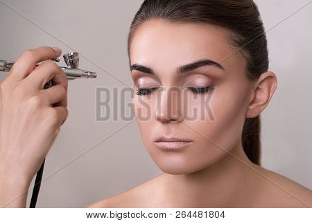 Visagist Making Makeup For Model With Aerograph, Isolated On Grey. Background With Hand Holding Aero