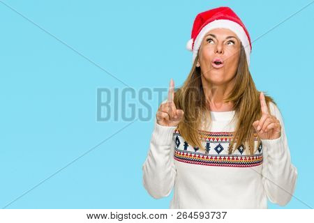 Middle age adult woman wearing winter sweater and chrismat hat over isolated background amazed and surprised looking up and pointing with fingers and raised arms.