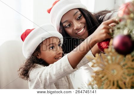 African American Mother And Daughter In Santa Claus Hats Decorating Christmass Tree Together At Home