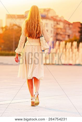 Fashion woman in autumn spring dress on city street and car. Back of female style of feminine fashionable girl model wallking hight heel fall outdoor. Color tone on shiny fountains sunlight background