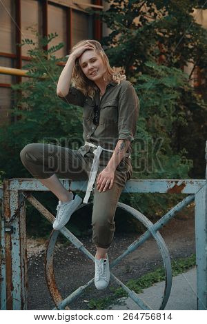 Stylish Girl Posing In The Street Sitting On A Steel Fence With Bent Legs. Urban Style. Street Fashi