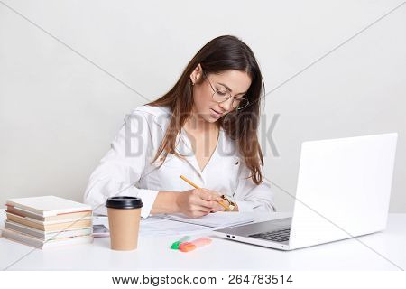 Female Blogger Wears White Shirt, Round Spectacles, Writes Notes In Notepad, Works On Laptop Compute
