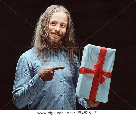 Happy Male With Long Hair And Beard In Blue Shirt Pointing On Gift Box In His Hand. Christmas Concep