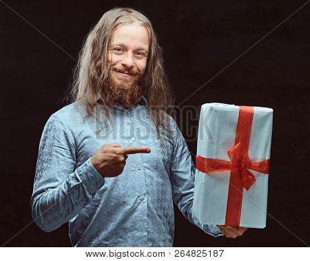 Happy Male With Long Hair And Beard In Blue Shirt Pointing On Gift Box In His Hand. Christmas Concep