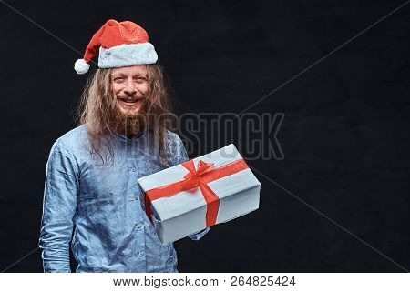 Happy Male With Long Hair And Beard In Blue Shirt And Red Santa Hat Holds Gift Box.