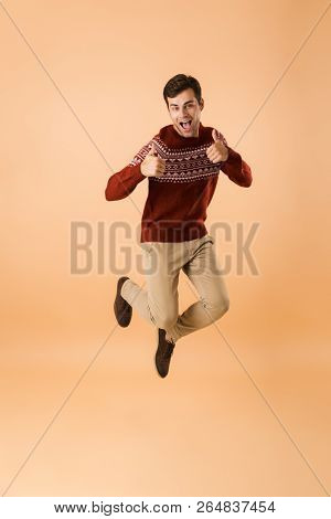Full length image of cheerful man 20s with bristle wearing knitted sweater jumping and rejoicing isolated over beige background