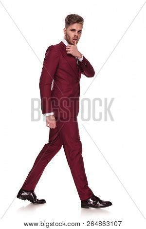 side view of sensual stylish man touching his lip while walking on white background, full length picture