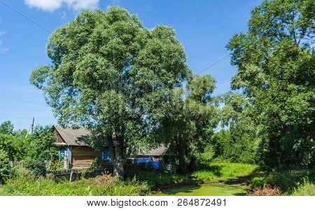 Wooden Village House Among The Gardens Of Green Trees Against The Background Of Red And Blue Sky And