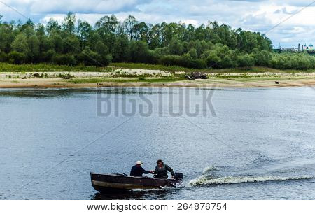 View Of The Pripyat River: Fishermen Move In A Motorboat Through The Water Against A Background Of G