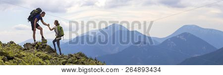 Young Tourists With Backpacks, Athletic Boy Helps Slim Girl To Clime Rocky Mountain Top Against Brig