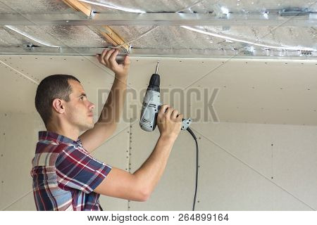 Young Man In Usual Clothing And Work Gloves Fixing Drywall Suspended Ceiling To Metal Frame Using El