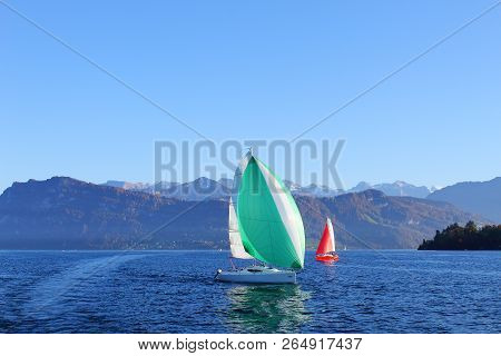 Active Rest On Clear Sunny Day. Yachts With Red And Green Sail On Lake Surrounded By Mountains.