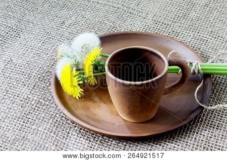 Ceramic Coffee Cup And Bouquet Of Dandelions On Round Clay Dish. Rough Homespun Background Of Jute.