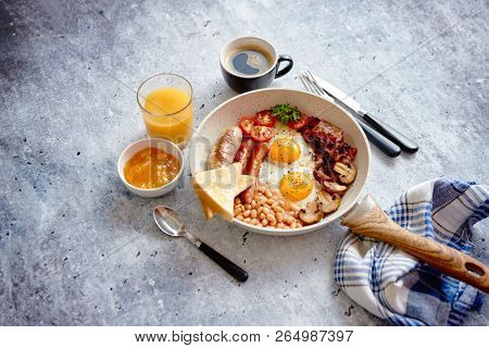 Full English Breakfast served in a pan. Fried eggs, beked beans, tomatoes, champignons, crispy bacon, sausages and toast. Placed on stone background. Top view with copy space.