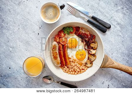Full English Breakfast served in a pan. Fried eggs, beked beans, tomatoes, champignons, crispy bacon, sausages and toast. Placed on stone background. Top view with copy space.