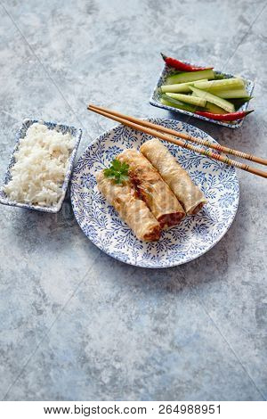 Orginal asian deep fried spring rolls placed on beautiful oriental style plate with chopstick, boiled rice, cucomber nad hot chilli peppers od side. Top view on stone background.