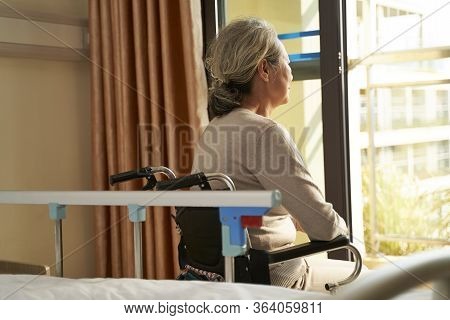 Rear View Of Senior Asian Woman Sitting In Wheel Chair In Nusing Home Or Hospital Ward Looking Out O
