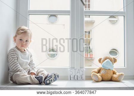 Child In Home Quarantine Playing At The Window With His Sick Teddy Bear Wearing A Medical Mask Again