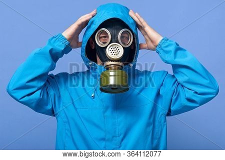 Horizontal Portrait Of Attentive Guy Putting On Hood, Wearing Gas Mask And Blue Jacket, Raising Hand