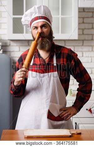 Bearded Chef Sticks A Rolling Pin In His Nose. Crazy Chef In A White Kitchen. Very Funny Photo.