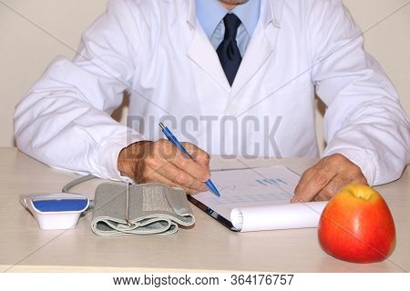 Close-up - The Doctor In A White Coat Sits At The Table And Eats A Healthy Apple.