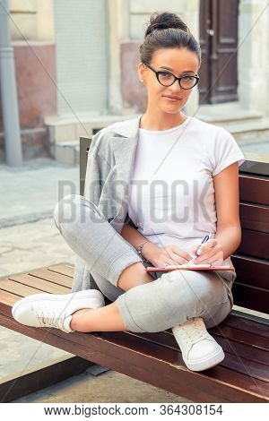 Smiling Young Woman With Glasses Is Making Notes In Notebook While Sitting On The Beanch Outdoors, L
