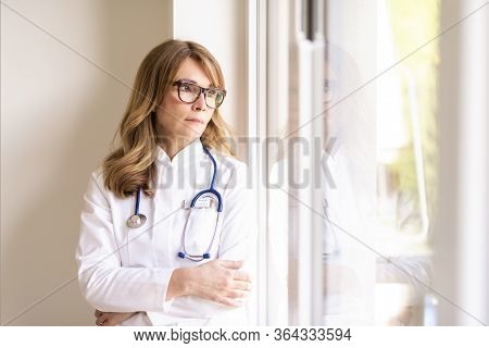Female Doctor Wearing Face Mask And Looking Careworn While Looking Out The Window During Coronavirus