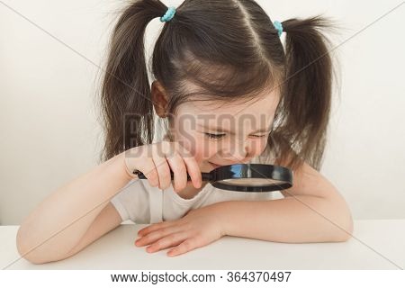 Little Girl Plays A Detective. A Child Examines Something Through A Magnifying Glass. A Girl With Tw