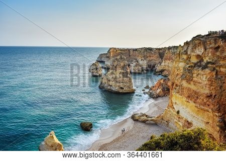 View From Above Of The Marinha Beach. Only One Couple Walks On The Sand. Orange Cliffs And Turquoise