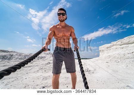 Fitness Athlete In A Cap And Sunglasses Training With Ropes Outdoor. Heavy Training In A Quarry. Int