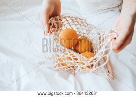 Woman Takes Oranges From A Net Eco Bag On A White Bed At Home.