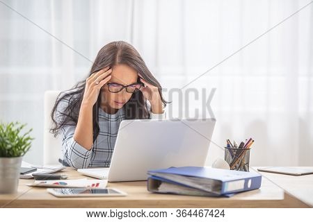Woman Having A Migrene Holding Head In Her Hands With Eyes Closed At The Desk, In Front Of The Compu