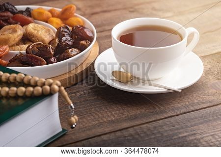 Closeup Iftar Concept, White Cup Of Tea, Plate Of Dried Fruit, Koran With Prayer Beads On Brown Wood