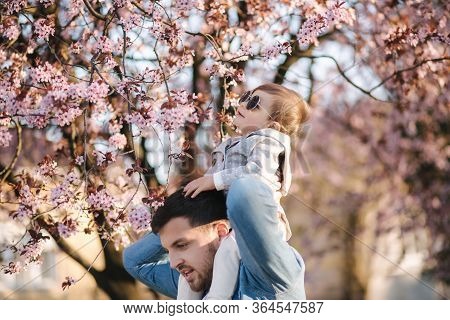 Adorable Little Daughter Sitting On Dads Neck And Smels Pink Flowers From Flowering Tree. Happy Fami
