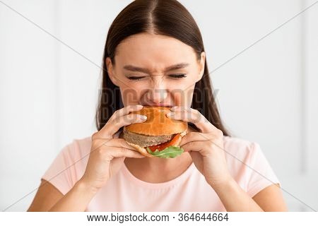 Cheat Meal. Hungry Girl Biting Burger Enjoying Eating Junk Food Having Diet Break Standing At Home.