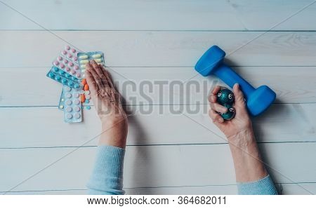 Senior Wrinkled Hand Pushing Back Pills And Thermometer, Dumb-bell On A Table. Another Old Woman Han